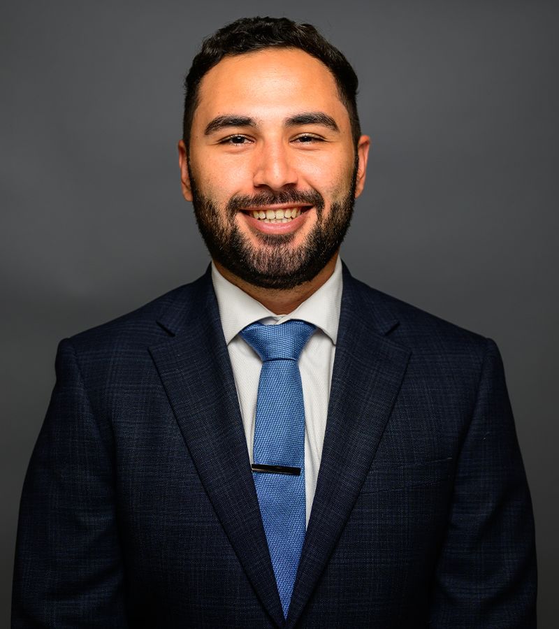 Giancarlo Pinto in a dark suit and blue tie against a gray background, professional headshot.