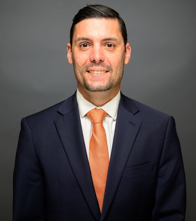 Ernesto A. Perez in suit with orange tie against gray background, professional portrait.