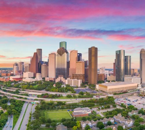 Houston skyline at sunset with colorful clouds, towering skyscrapers, and green parks in the foreground.