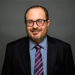 Stephen Ostrow in glasses and suit with a checkered shirt and striped tie, posing against a gray background.