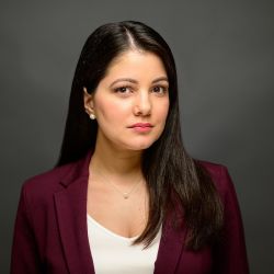 Professional woman in maroon blazer with long dark hair, serious expression, neutral background.
