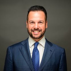 Alex Uriarte in a blue suit and tie on a gray background.