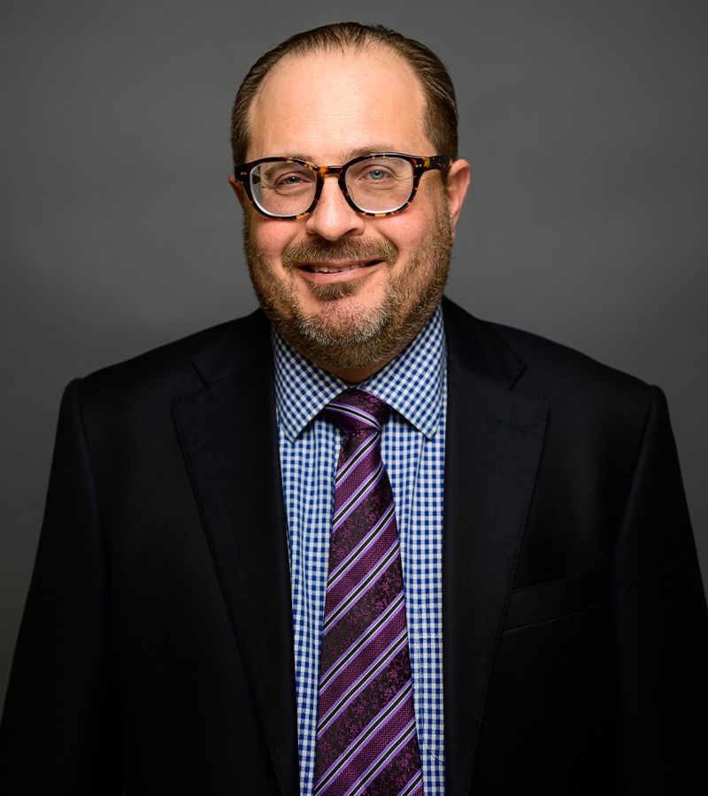 Stephen Ostrow in glasses and suit with a checkered shirt and striped tie, posing against a gray background.