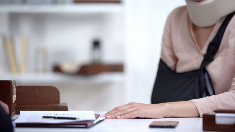 Injured woman with neck brace and arm sling at desk with medical negligence documents.