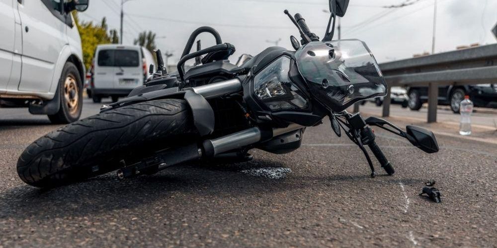 Motorcycle lies on its side in a road collision, front end damaged, with nearby vehicles and a bottle on the pavement nearby, suggesting an accident scene.