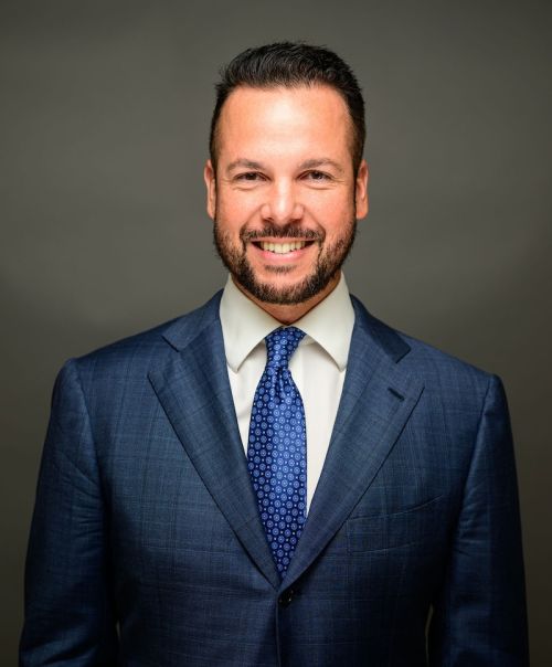 Alex Uriarte in a blue suit and tie on a gray background.