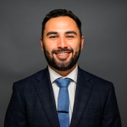 Giancarlo Pinto in a dark suit and blue tie against a gray background, professional headshot.