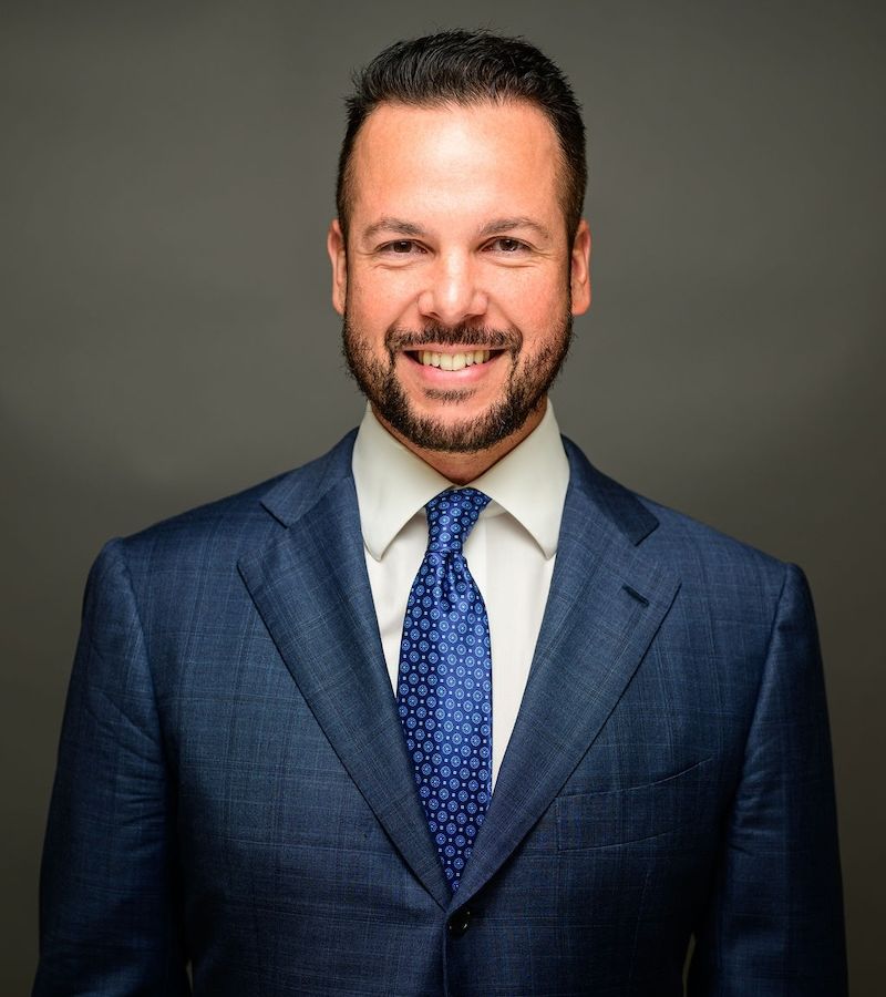 Alex Uriarte in a blue suit and tie on a gray background.