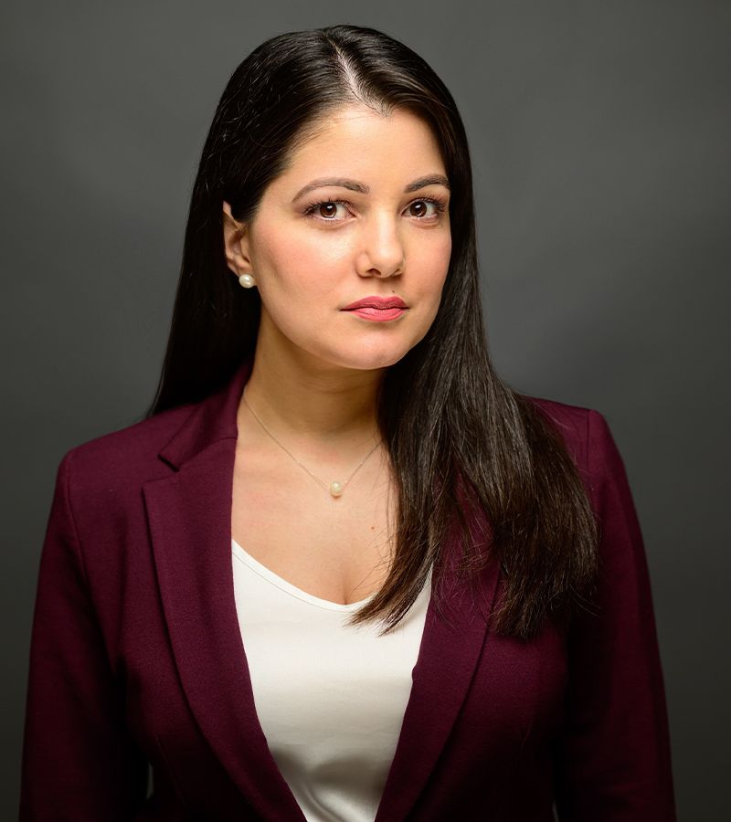 Professional woman in maroon blazer with long dark hair, serious expression, neutral background.
