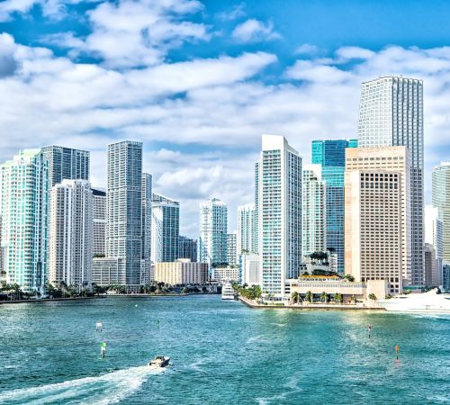 Skyline of a modern coastal city with high-rise buildings and a boat navigating the waterfront on a sunny day.