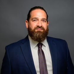 Man in a blue suit with a patterned tie, sporting a full beard, against a gray background.