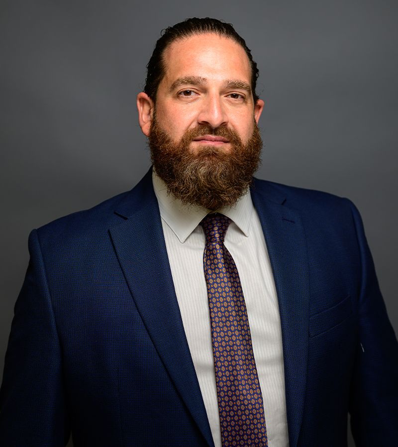 Man in a blue suit with a patterned tie, sporting a full beard, against a gray background.