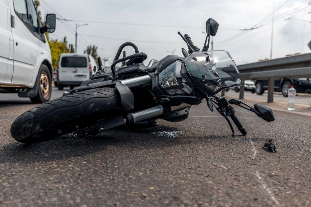 Motorcycle lies on its side in a road collision, front end damaged, with nearby vehicles and a bottle on the pavement nearby, suggesting an accident scene.
