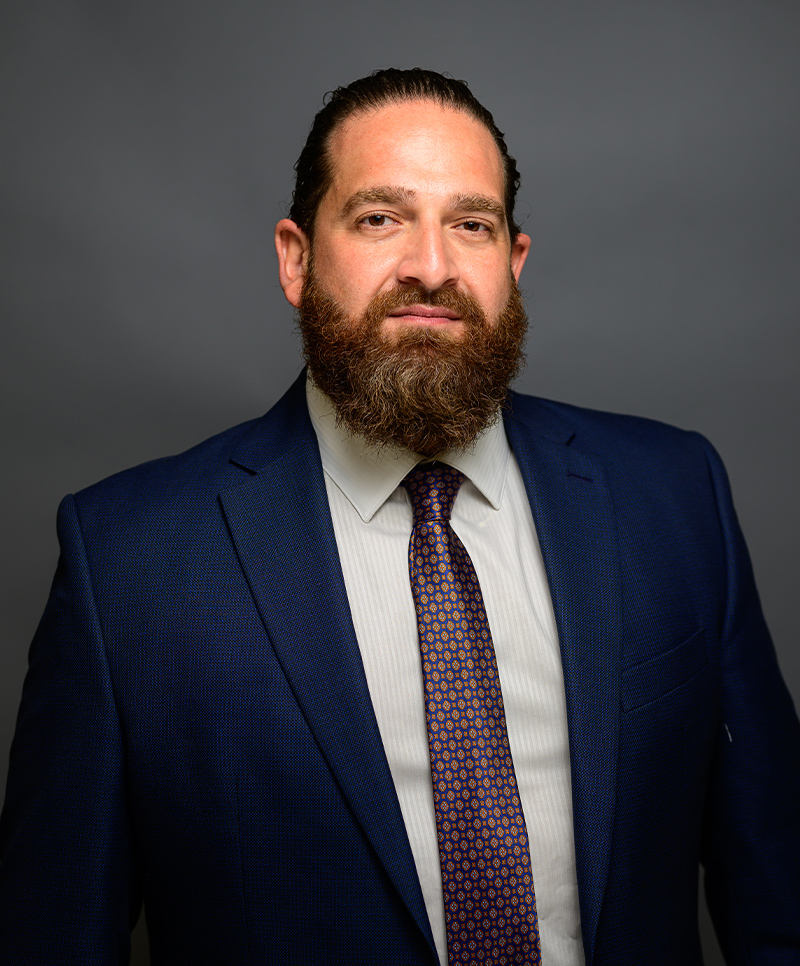 Man in a blue suit with a patterned tie, sporting a full beard, against a gray background.