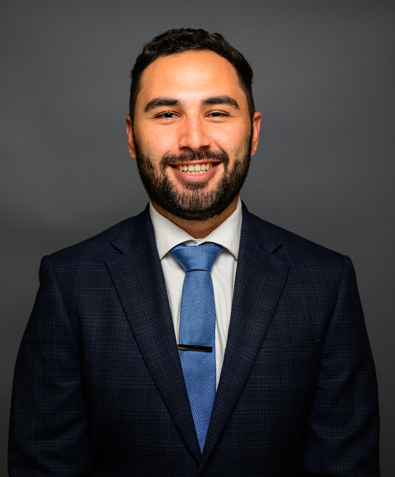 Giancarlo Pinto in a dark suit and blue tie against a gray background, professional headshot.