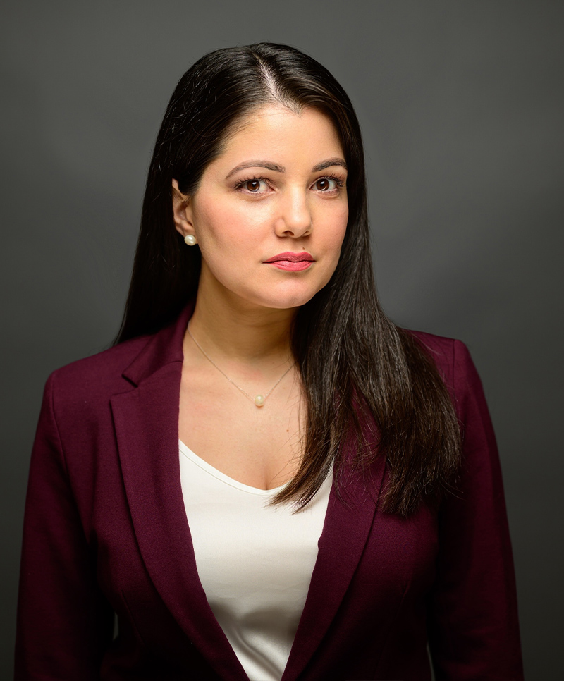 Professional woman in maroon blazer with long dark hair, serious expression, neutral background.